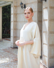 A woman wearing a flowing cream-colored kaftan dress with wide sleeves, standing in front of a stone building entrance.