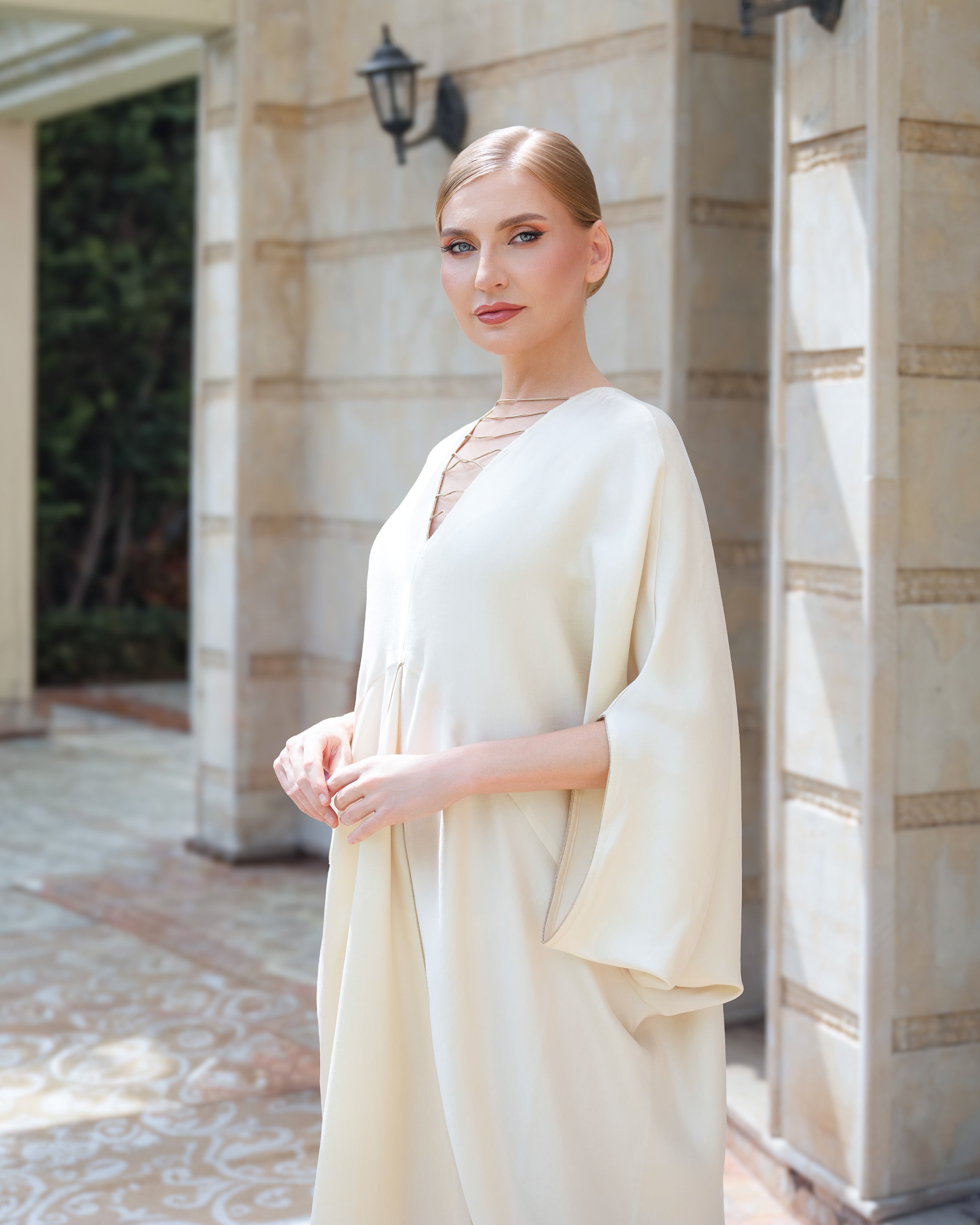A woman wearing a flowing cream-colored kaftan dress with wide sleeves, standing in front of a stone building entrance.