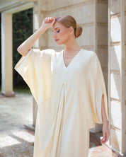 A woman wearing a flowing cream-colored kaftan dress with wide draped sleeves, standing in a sunlit outdoor corridor with stone columns.