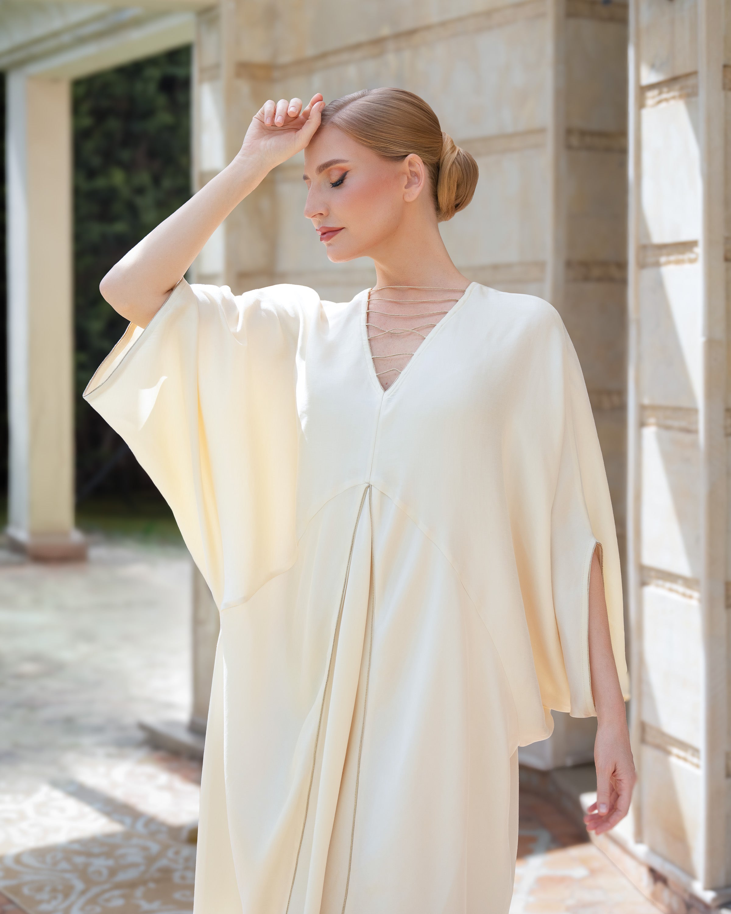 A woman wearing a flowing cream-colored kaftan dress with wide draped sleeves, standing in a sunlit outdoor corridor with stone columns.