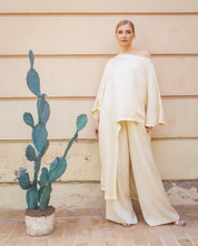 A woman wearing a flowing, off-the-shoulder cream-colored dress or caftan with wide draped sleeves, standing against a beige paneled wall.