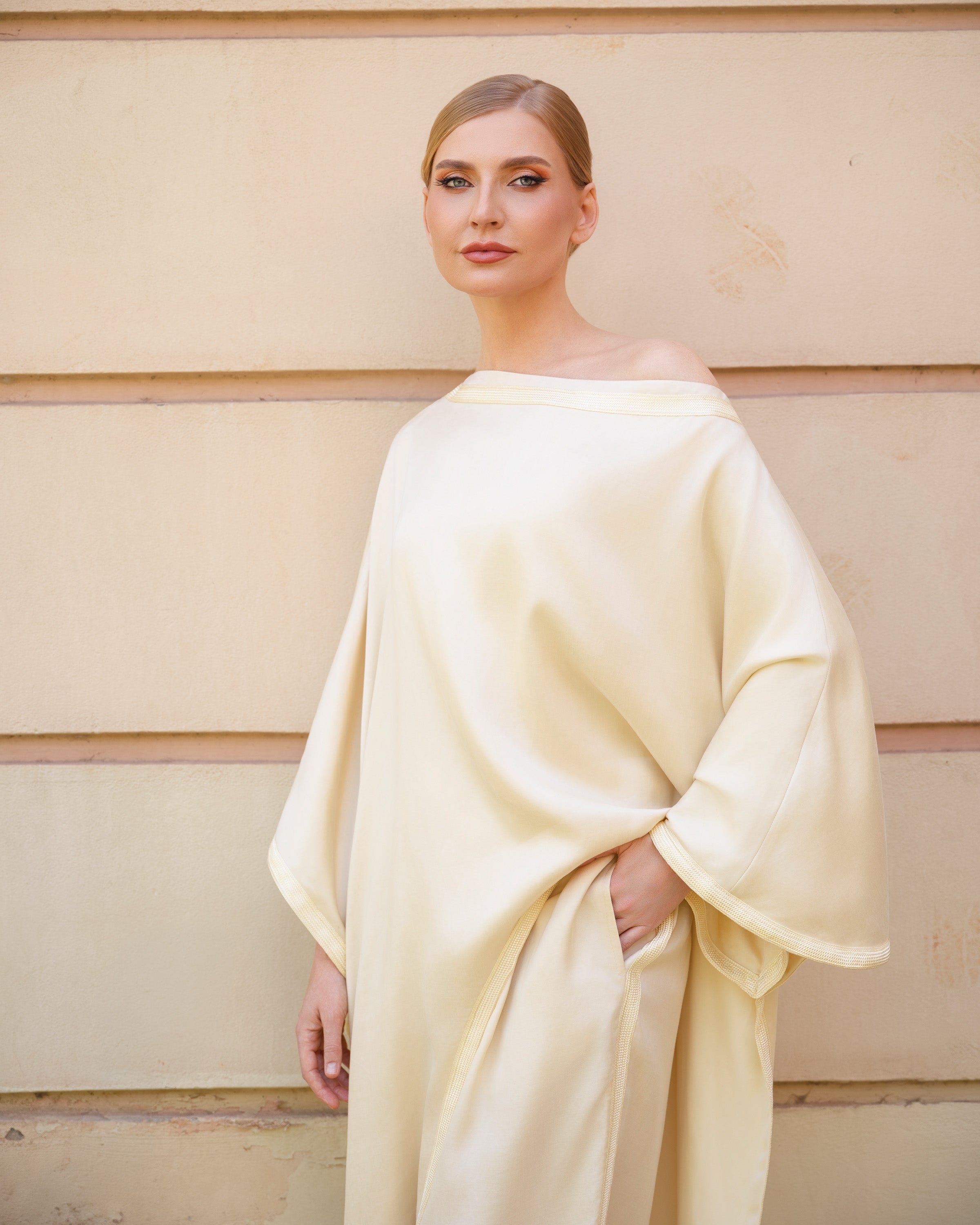 A woman wearing a flowing, off-the-shoulder cream-colored dress or caftan with wide draped sleeves, standing against a beige paneled wall.