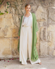 A woman stands against a rustic stone wall wearing a sage green open-front robe draped over a cream-colored top and matching high-waisted trousers.
