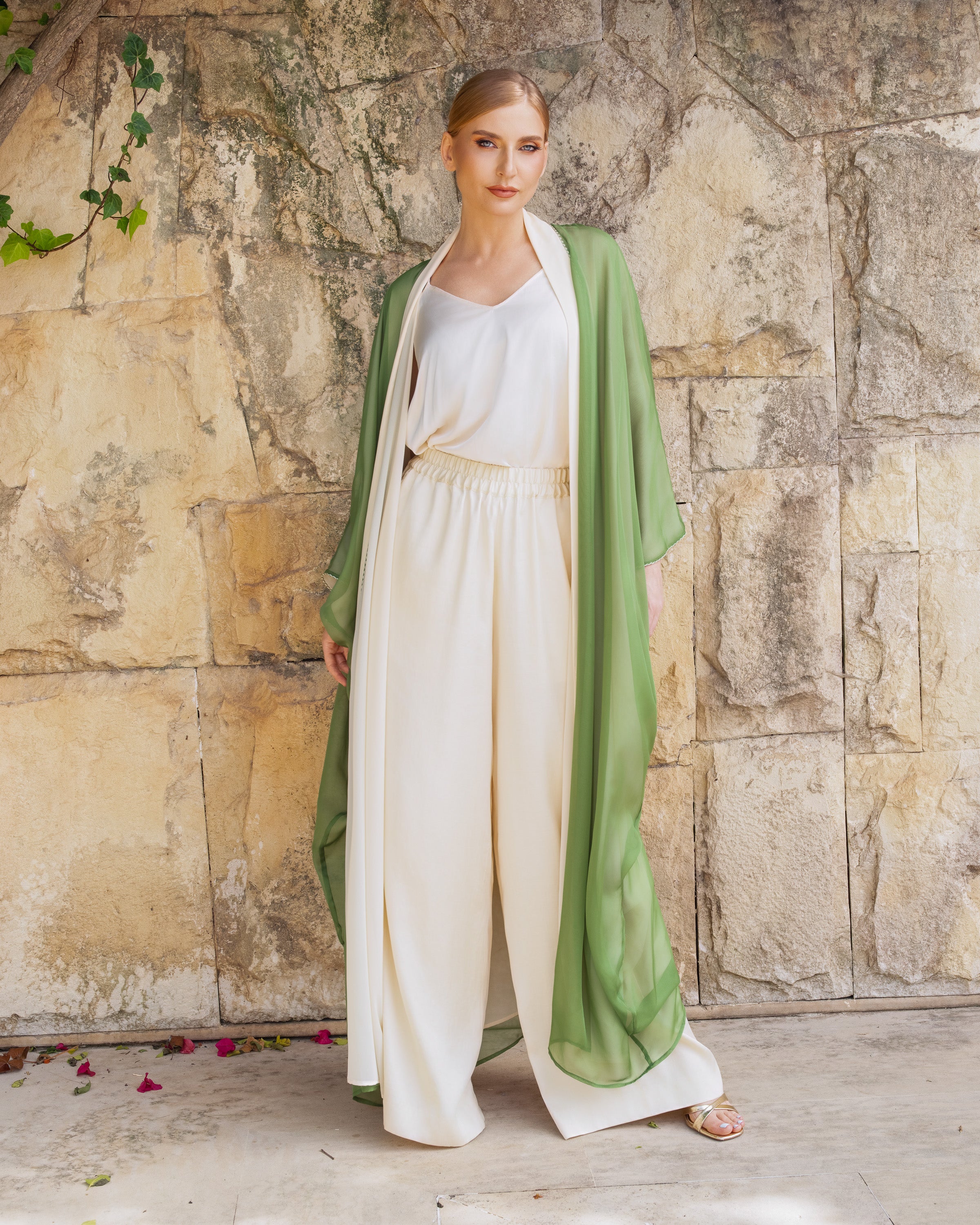 A woman stands against a rustic stone wall wearing a sage green open-front robe draped over a cream-colored top and matching high-waisted trousers.