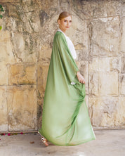 A woman wearing a flowing sage green kaftan dress with a white inner top, standing in front of a rustic stone wall.