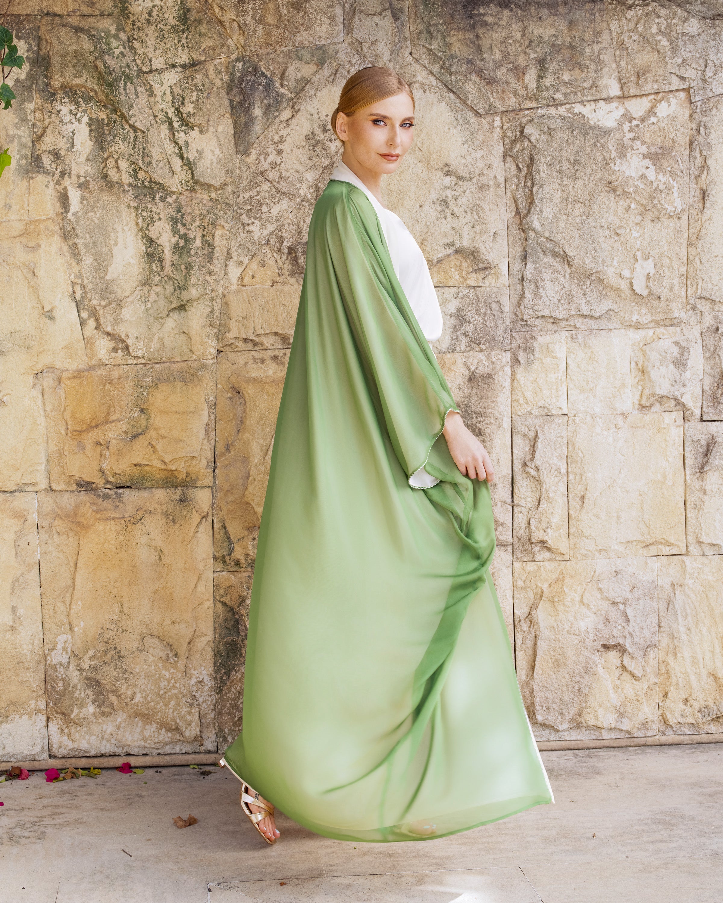 A woman wearing a flowing sage green kaftan dress with a white inner top, standing in front of a rustic stone wall.