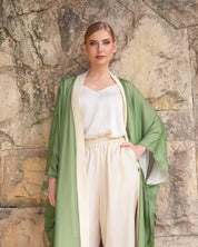 A woman stands against a rustic stone wall wearing a sage green open-front robe draped over a cream-colored top and matching high-waisted trousers.