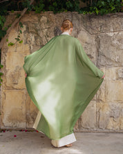 A woman viewed from behind wearing a flowing sage green wrap or robe over cream-colored garments, standing in front of a rustic stone wall with green ivy.