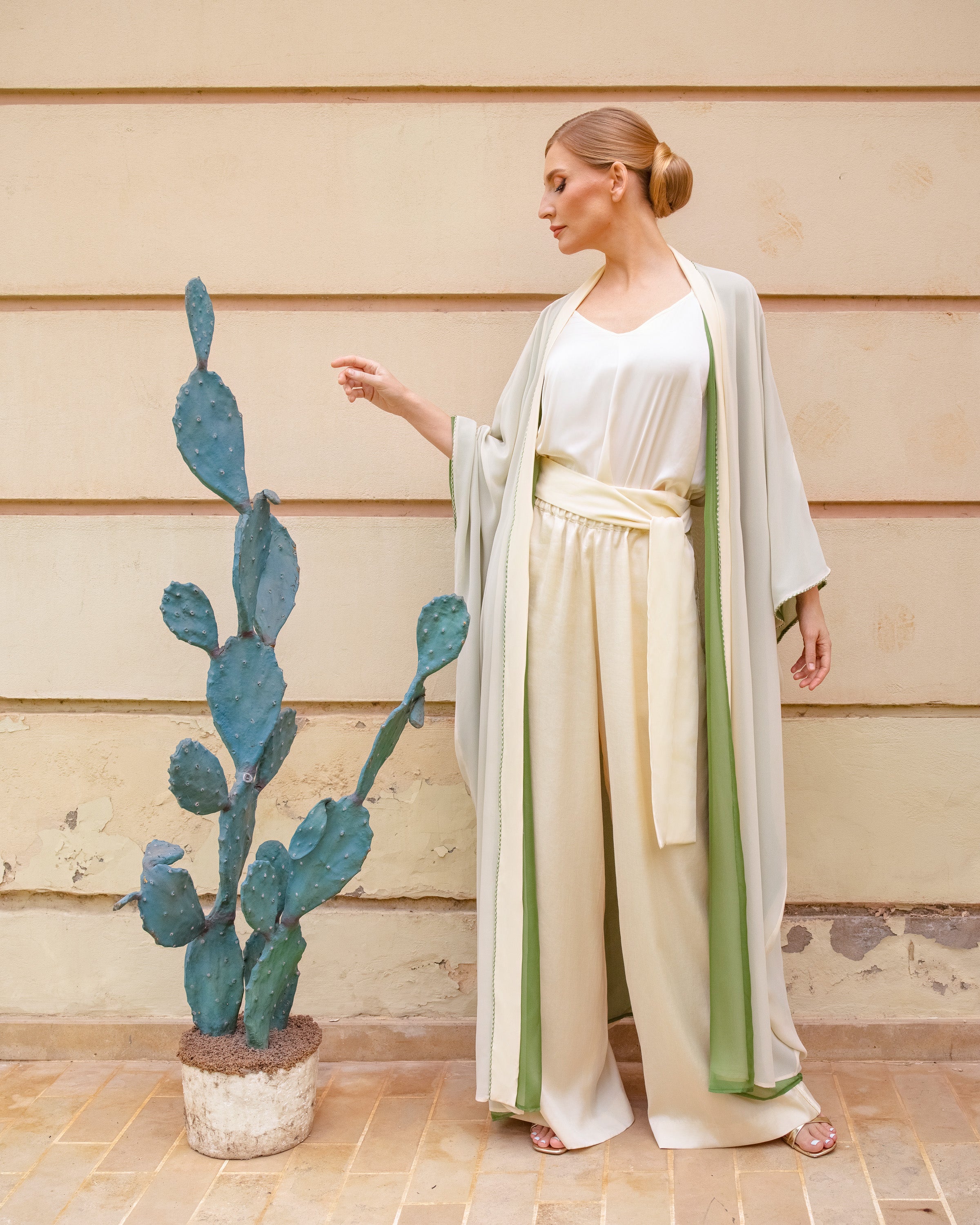 A woman wearing a cream and sage green draped kimono-style outfit stands beside a potted cactus against a beige wooden wall.