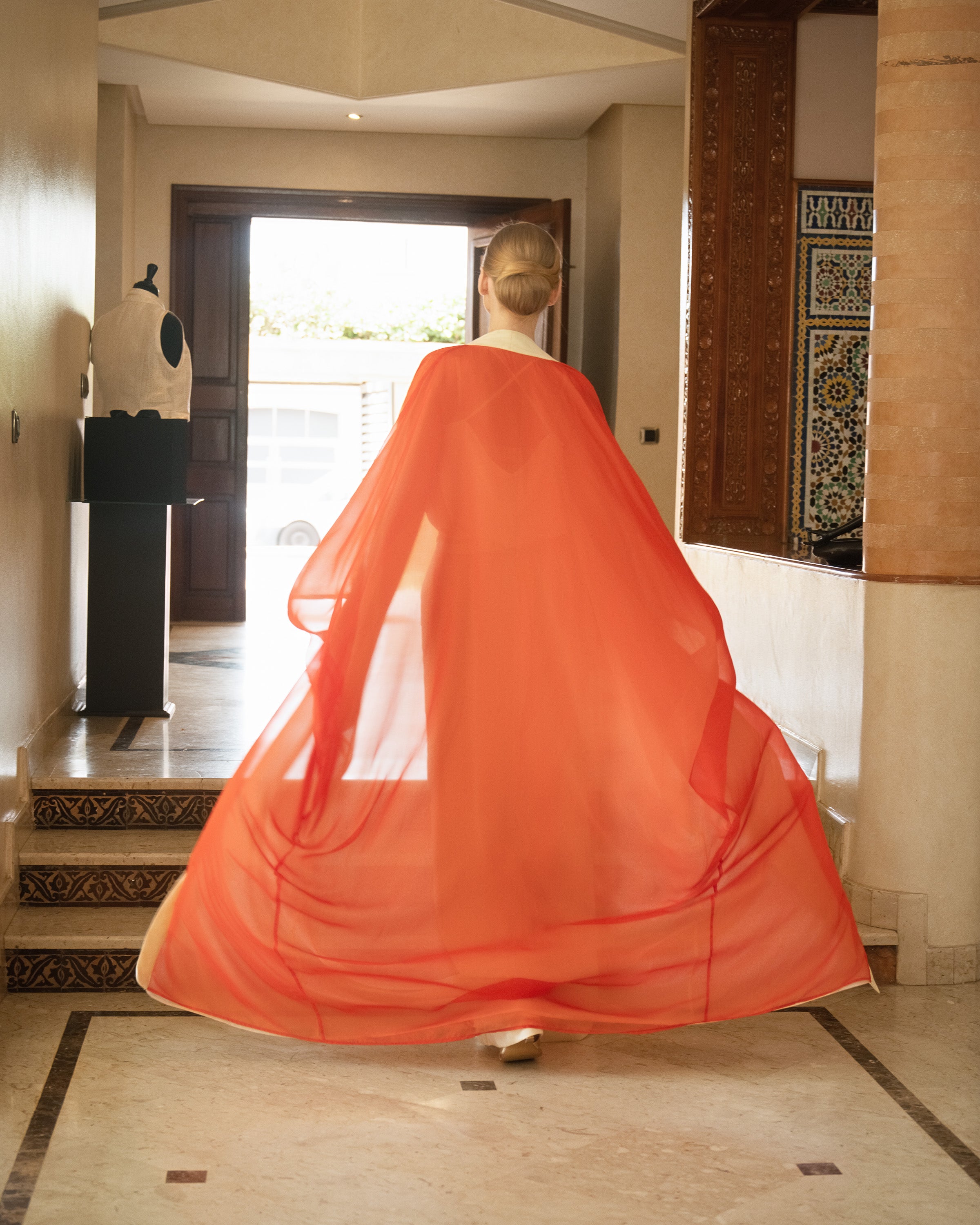 A woman viewed from behind wearing a flowing sheer orange-red shawl or cape over a white dress, standing in a tiled indoor hallway with a doorway behind her.