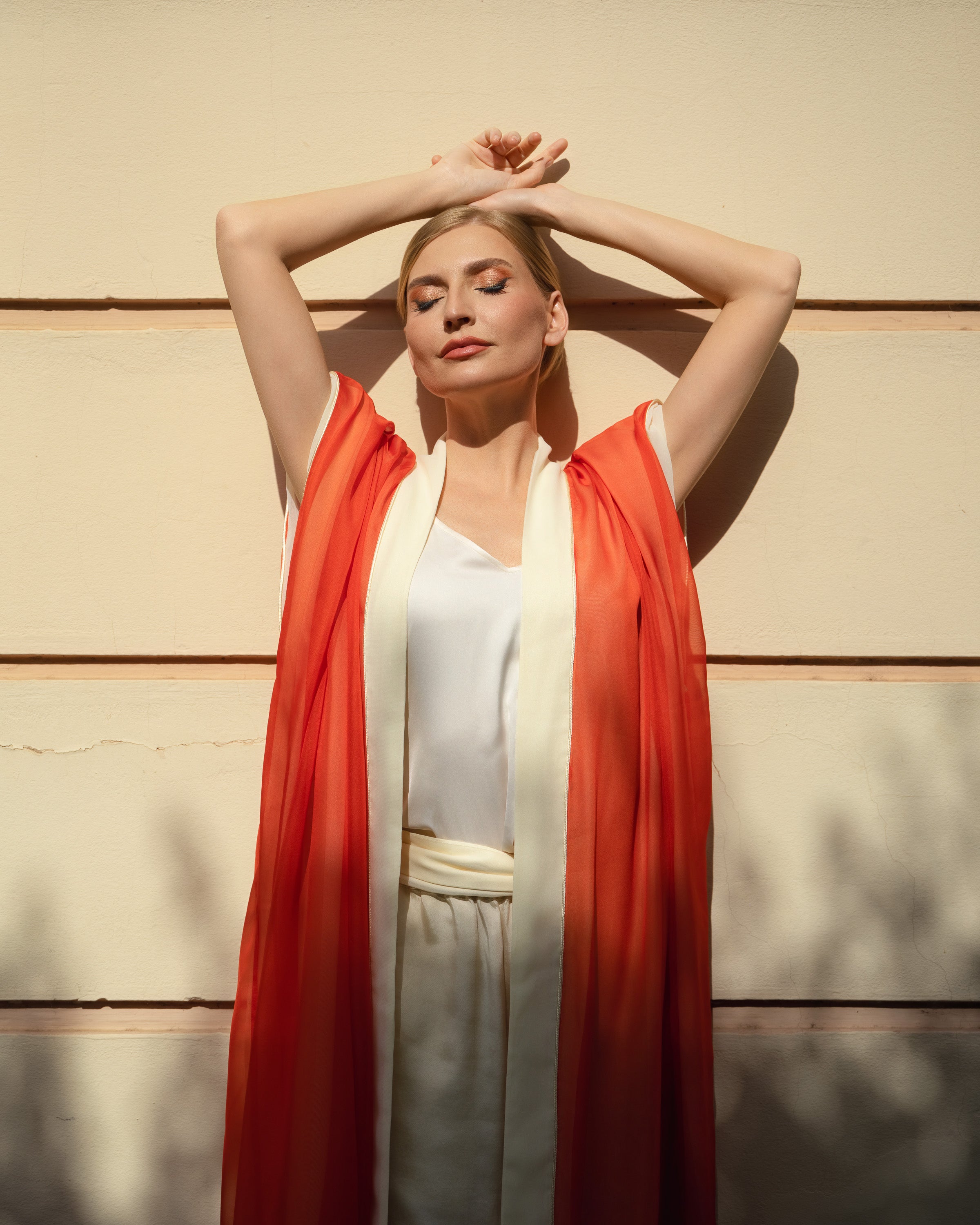 A woman posing outdoors against a beige wall, wearing a white sleeveless top and light beige trousers, with a flowing orange-red open-front drape or kimono layered over her shoulders.
