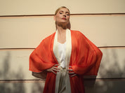 A woman posing outdoors against a beige wall, wearing a white sleeveless top and light beige trousers, with a flowing orange-red open-front drape or kimono layered over her shoulders.