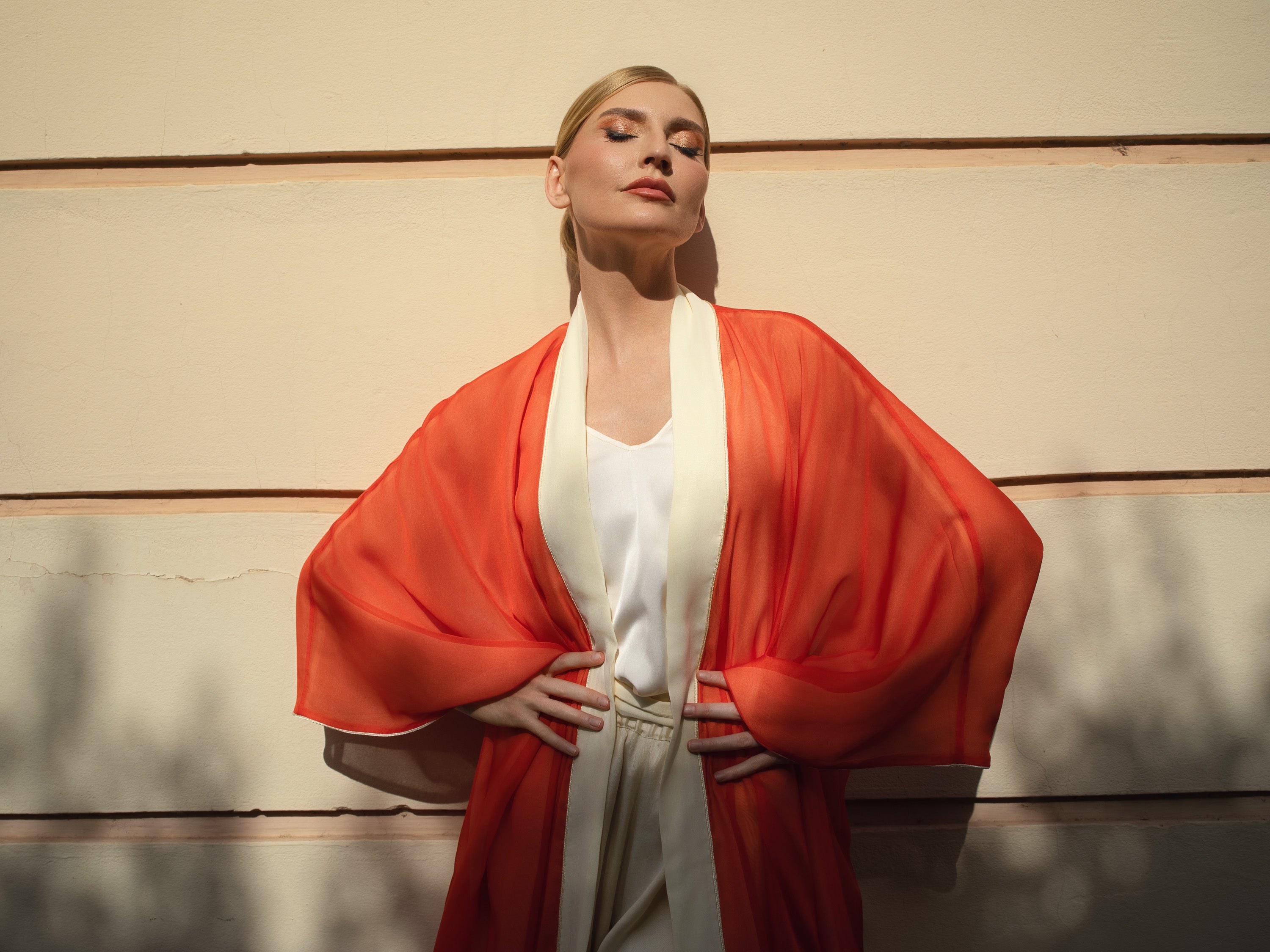 A woman posing outdoors against a beige wall, wearing a white sleeveless top and light beige trousers, with a flowing orange-red open-front drape or kimono layered over her shoulders.