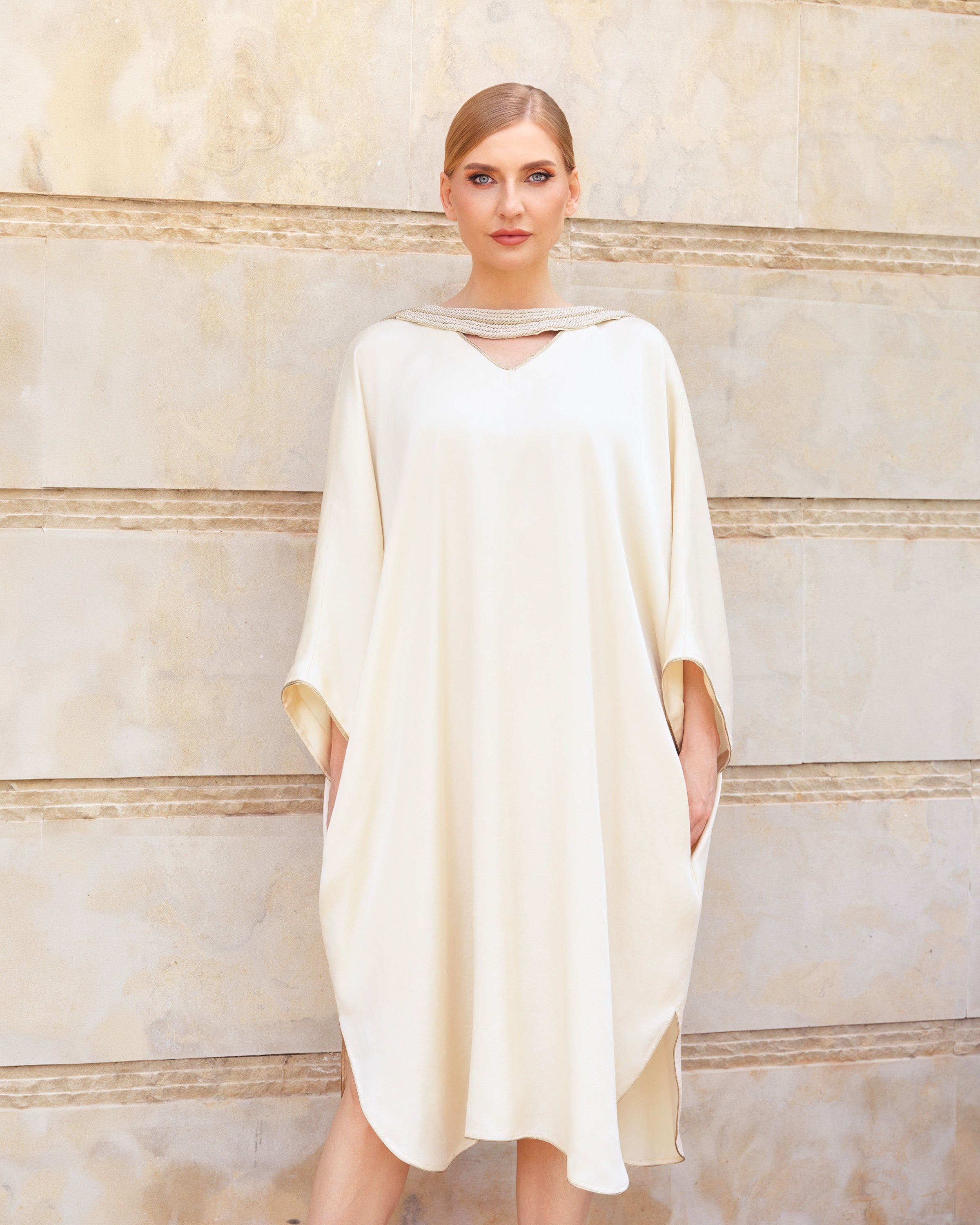 A woman modeling a cream-colored, loose-fitting kaftan dress with a keyhole cutout at the neckline, standing against a beige stone wall.