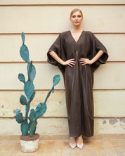 A woman stands against a beige paneled wall wearing a loose, long brown kaftan dress with wide draped sleeves and a wrap-style front. A potted cactus sits to her left.