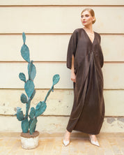 A woman wearing a dark brown, loose-fitting kaftan dress with three-quarter sleeves and a deep V-neckline, standing against a beige wooden wall next to a potted cactus.