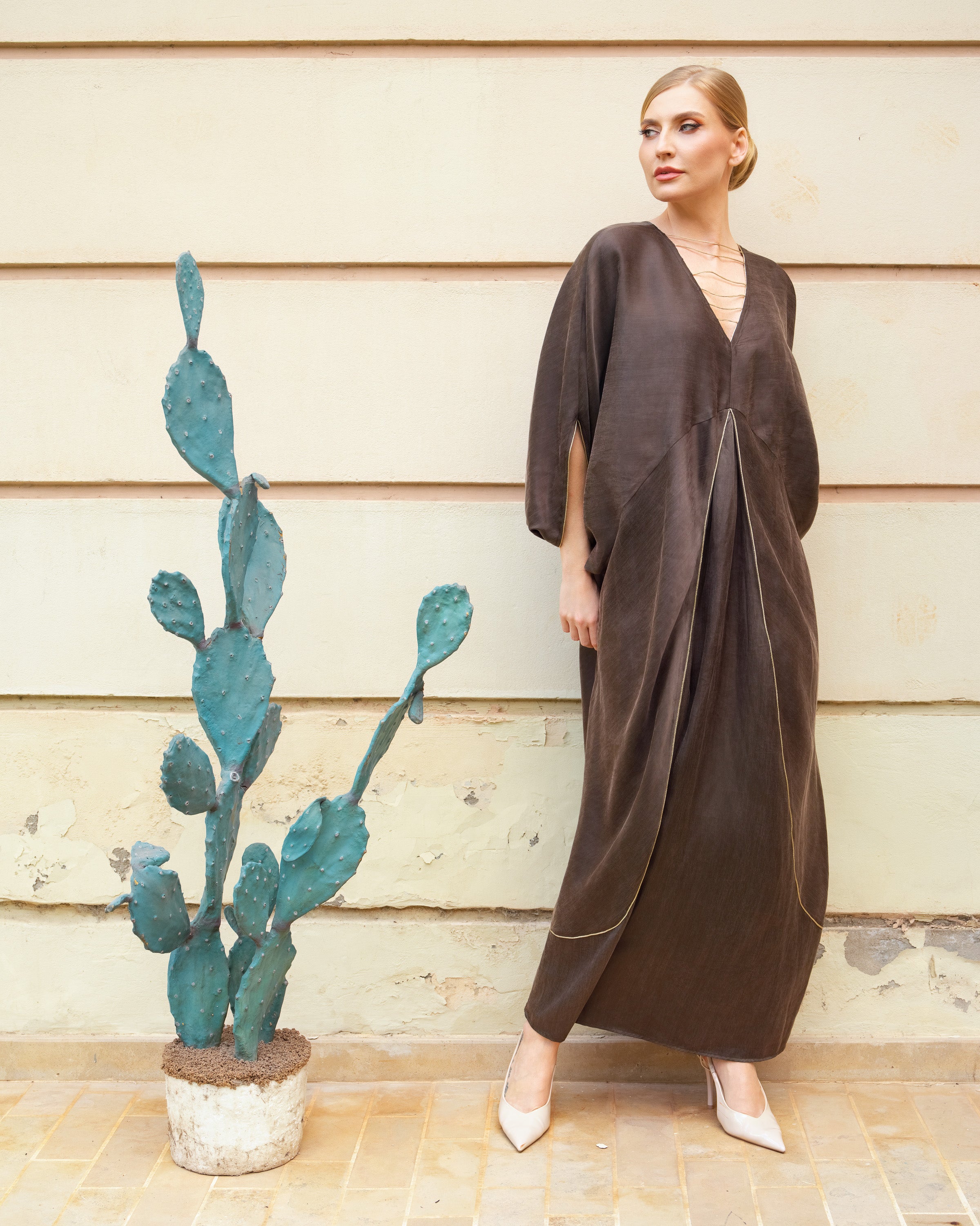 A woman wearing a dark brown, loose-fitting kaftan dress with three-quarter sleeves and a deep V-neckline, standing against a beige wooden wall next to a potted cactus.