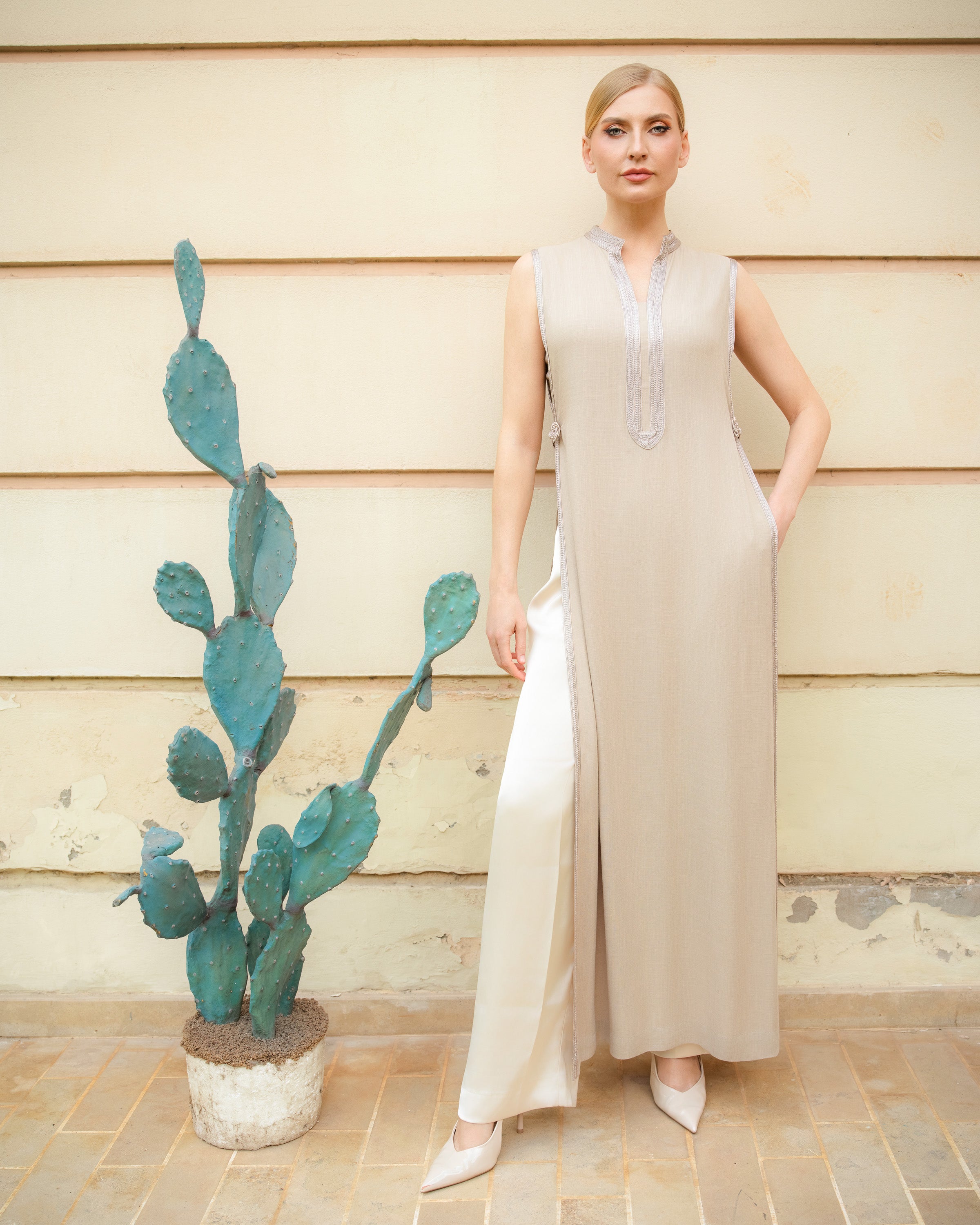 A woman models a sleeveless, floor-length beige maxi dress with a high neckline and silver embroidery down the front placket. She is standing next to a potted cactus in front of a beige wooden wall.