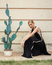A woman wearing a black sleeveless dress with beige trim sits on a tiled ledge against a beige wall, next to a potted cactus.