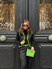 A woman with brown hair and sunglasses posing in front of ornate black doors, wearing a black outfit with striking neon green trim and holding a matching bright green quilted clutch.