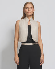 Woman wearing a beige vest over a black top and pants on a light gray background