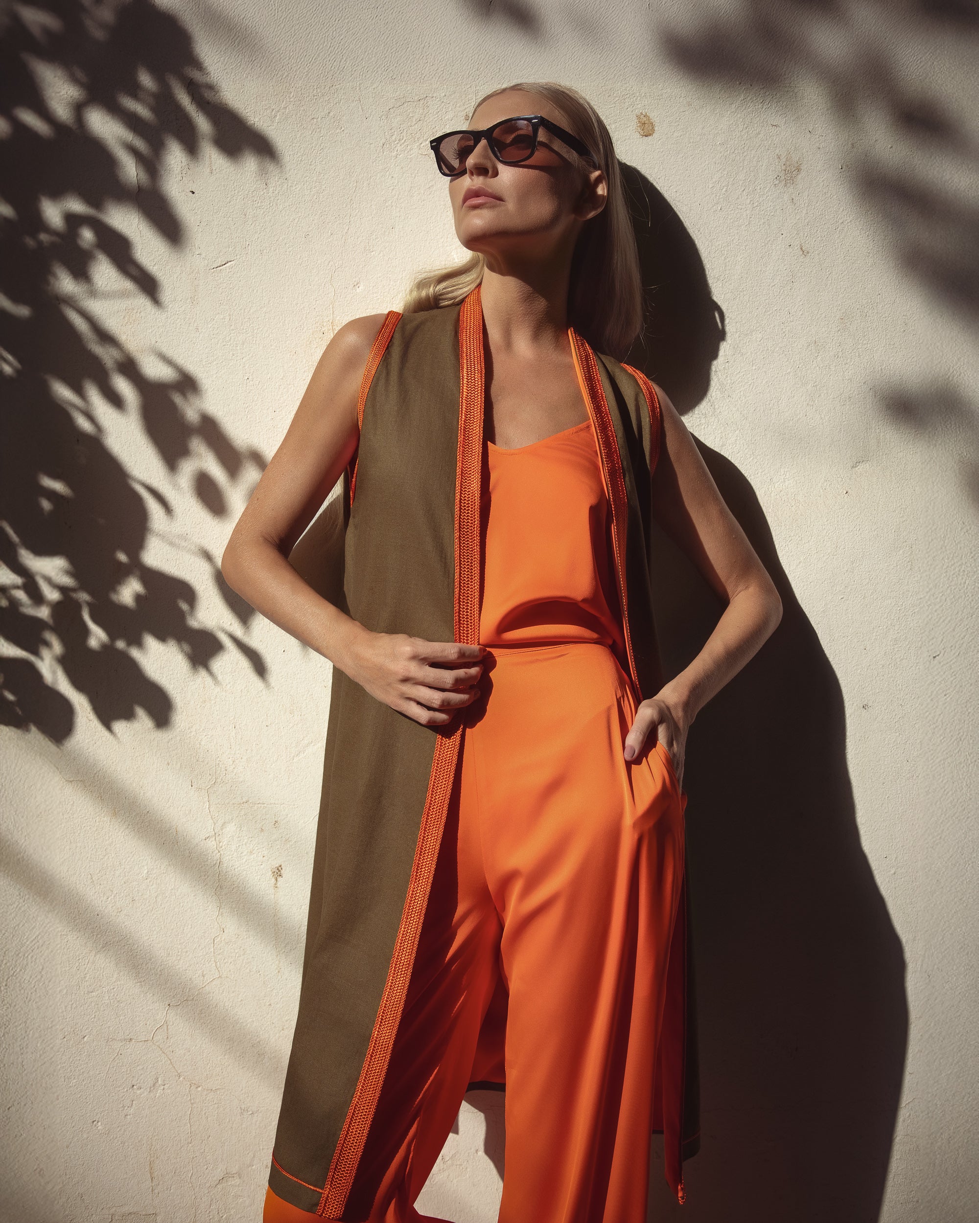 Woman in an orange outfit with a green vest standing against a textured wall.