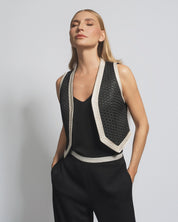 Woman wearing a black sleeveless top and pants with a textured vest against a plain background