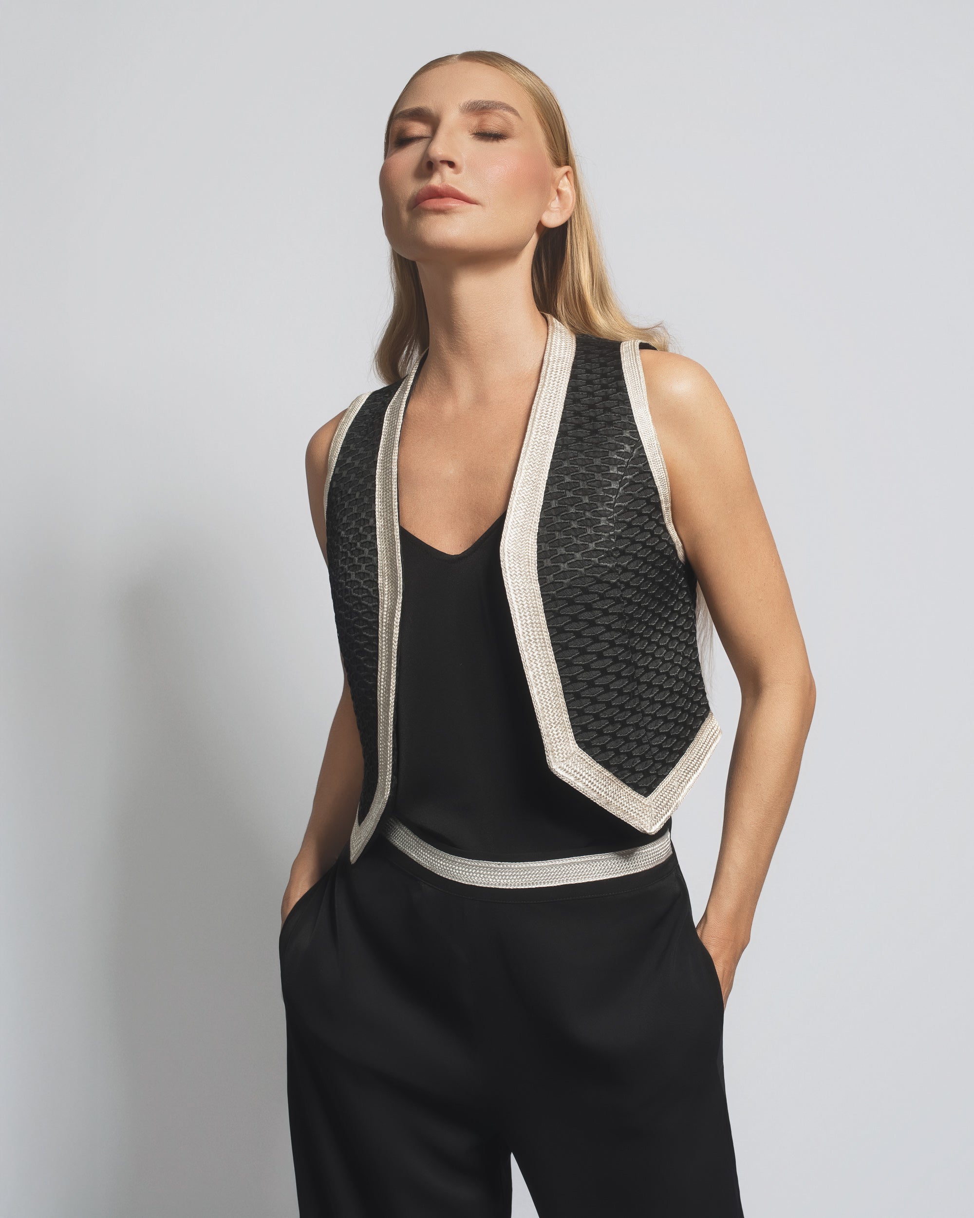 Woman wearing a black sleeveless top and pants with a textured vest against a plain background