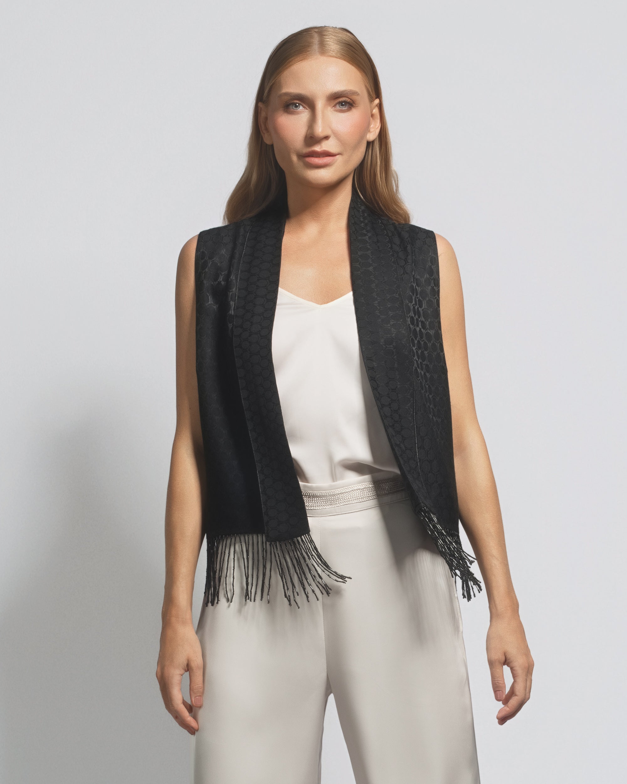 Woman wearing a black fringe vest over a white top and pants on a light gray background