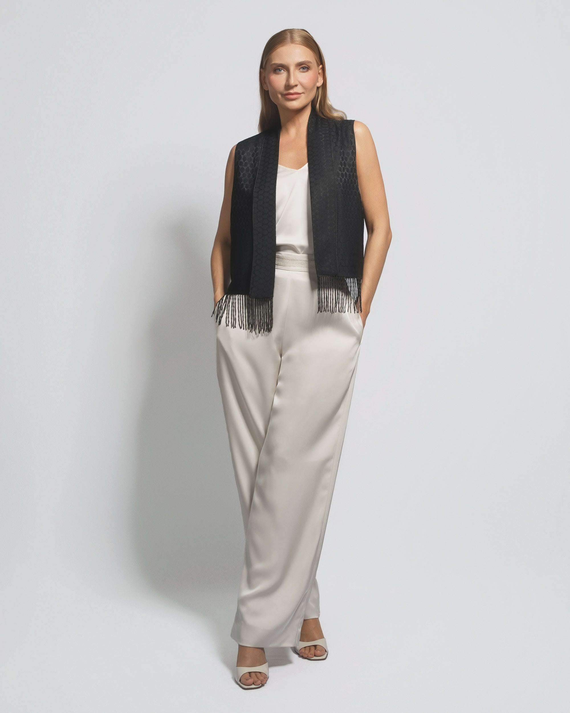 Woman wearing a black vest with fringe and white pants on a light gray background