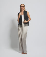 Woman wearing a black sleeveless top with fringe and beige pants on a plain background