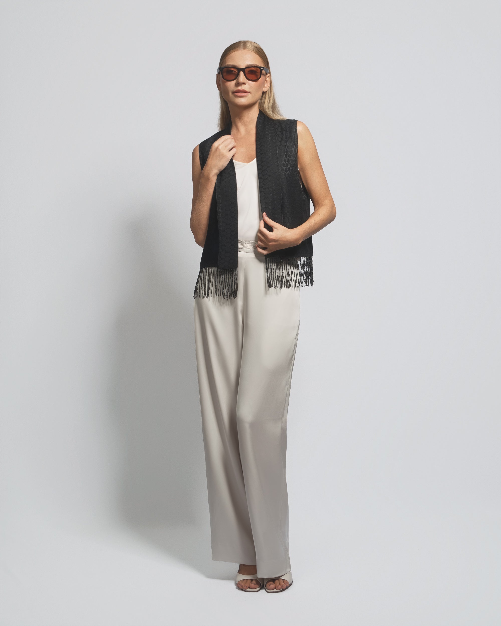 Woman wearing a black sleeveless top with fringe and beige pants on a plain background