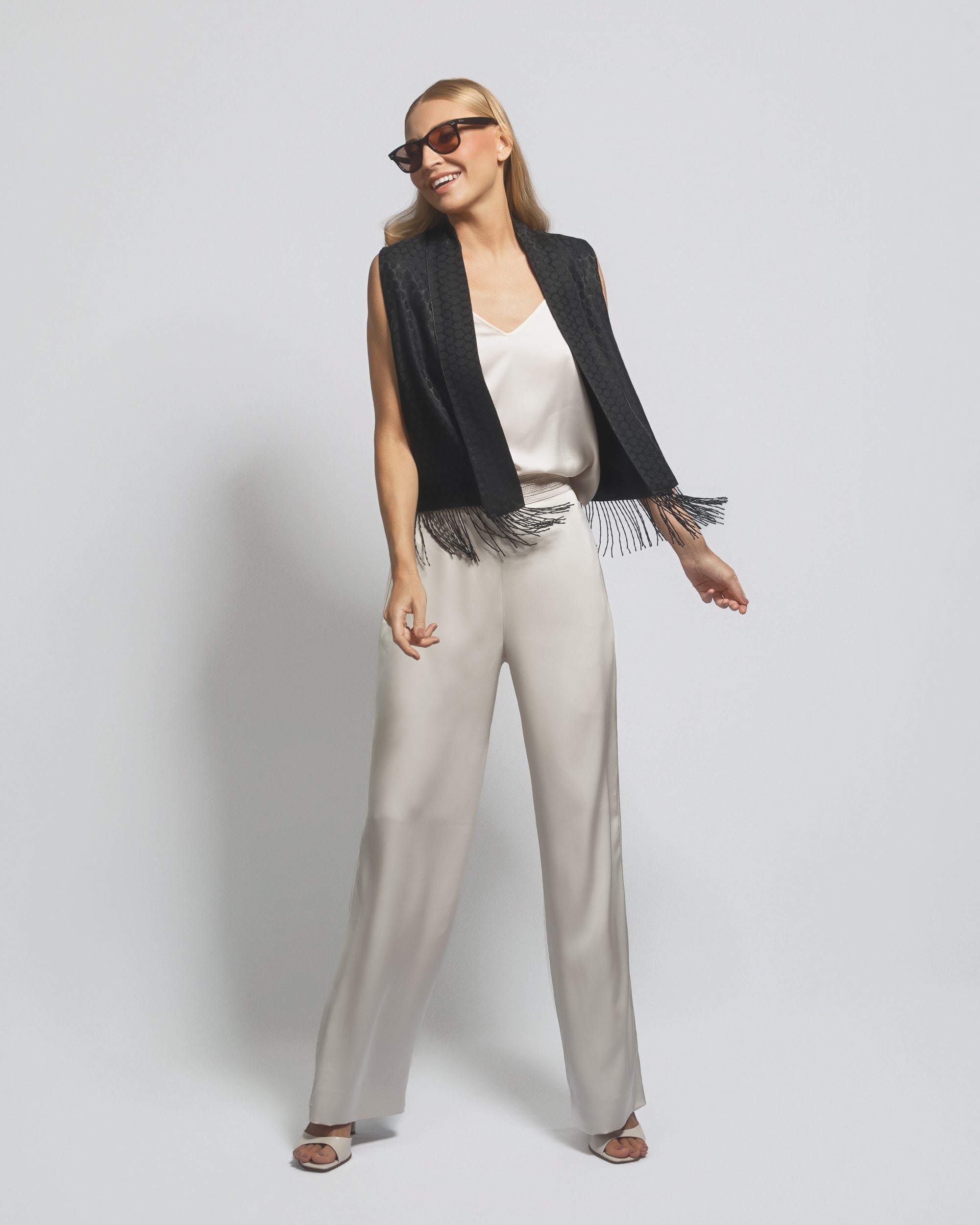 Woman wearing a black vest, white top, and beige pants on a plain background