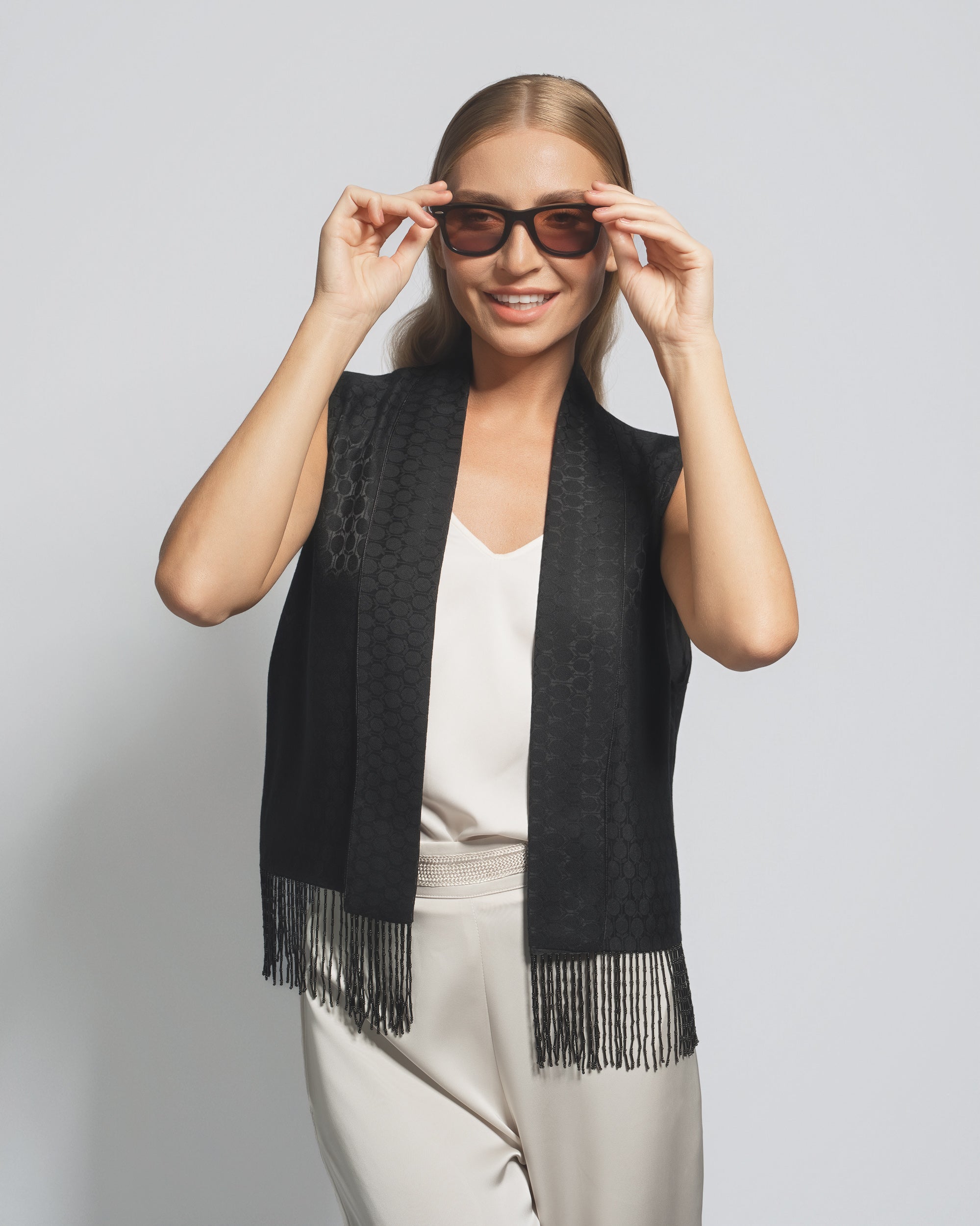 Woman wearing a black scarf with fringe, sunglasses, and a white top against a light gray background