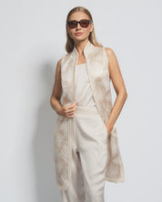 Woman wearing a beige sleeveless top with lace details and white pants against a light gray background.