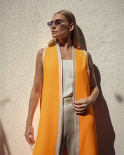 Woman wearing a bright orange vest over a white top and pants against a beige wall.