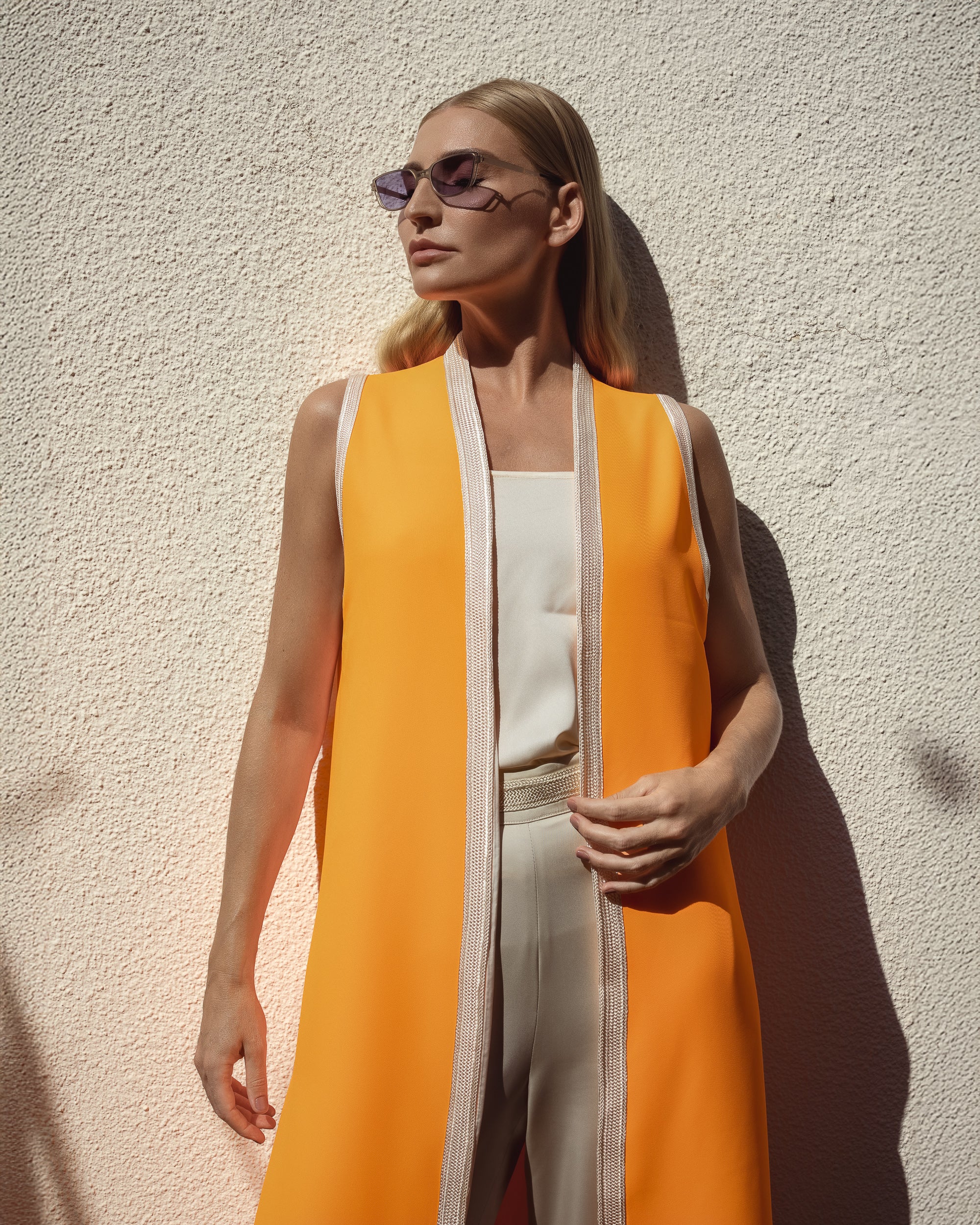 Woman wearing a bright orange vest over a white top and pants against a beige wall.