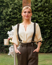 A woman stands outdoors in front of a green hedge, wearing a cream blouse with dark suspenders and dark brown trousers. She is standing next to a small glass table with white flowers.
