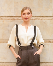 A woman wearing a cream-colored blouse with black suspenders and dark brown trousers, standing against a beige stone wall.