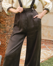 A woman wearing dark brown wide-leg trousers with black suspenders, paired with a cream blouse, standing outdoors next to a brick column.