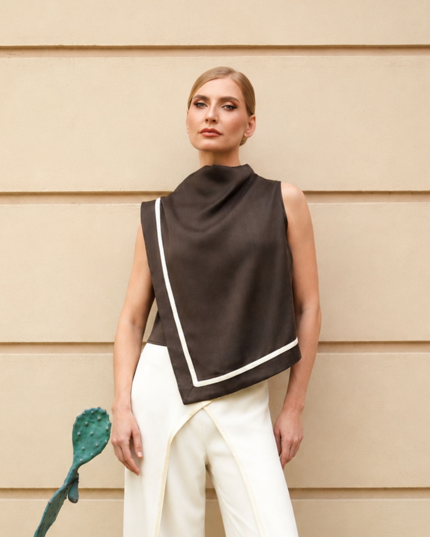 A woman stands against a beige paneled wall wearing a dark brown sleeveless wrap-style top with white contrast trim and white pants. A green cactus-shaped accessory is partially visible at the lower left.