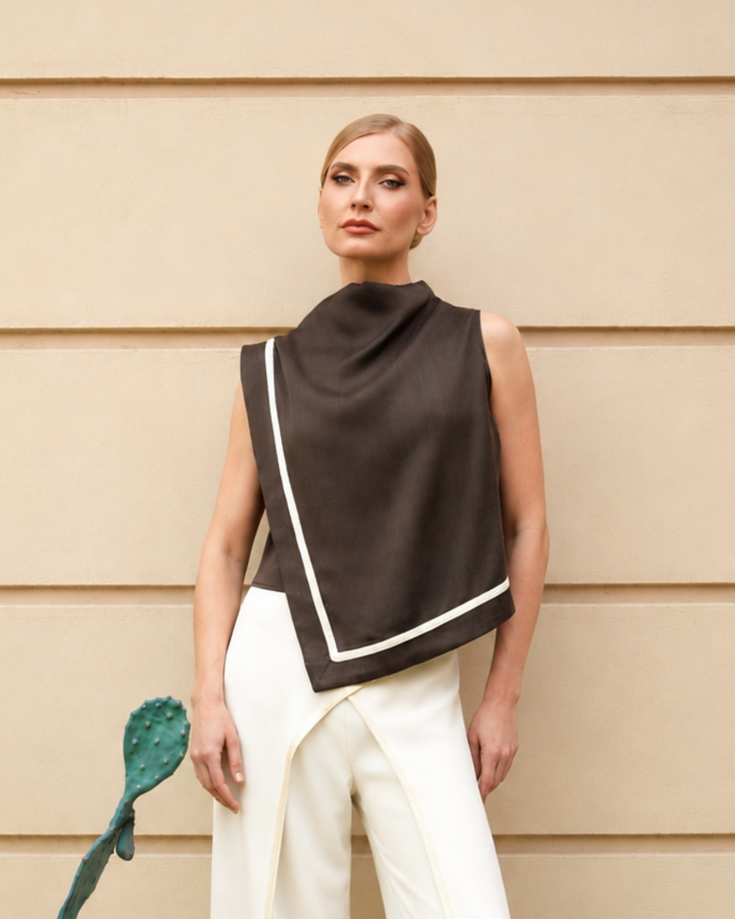 A woman stands against a beige paneled wall wearing a dark brown sleeveless wrap-style top with white contrast trim and white pants. A green cactus-shaped accessory is partially visible at the lower left.