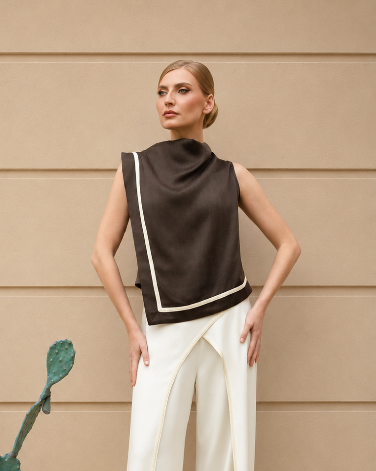 A woman models a dark brown sleeveless wrap-style top with cream-colored piping, paired with white draped trousers, set against a beige paneled wall.