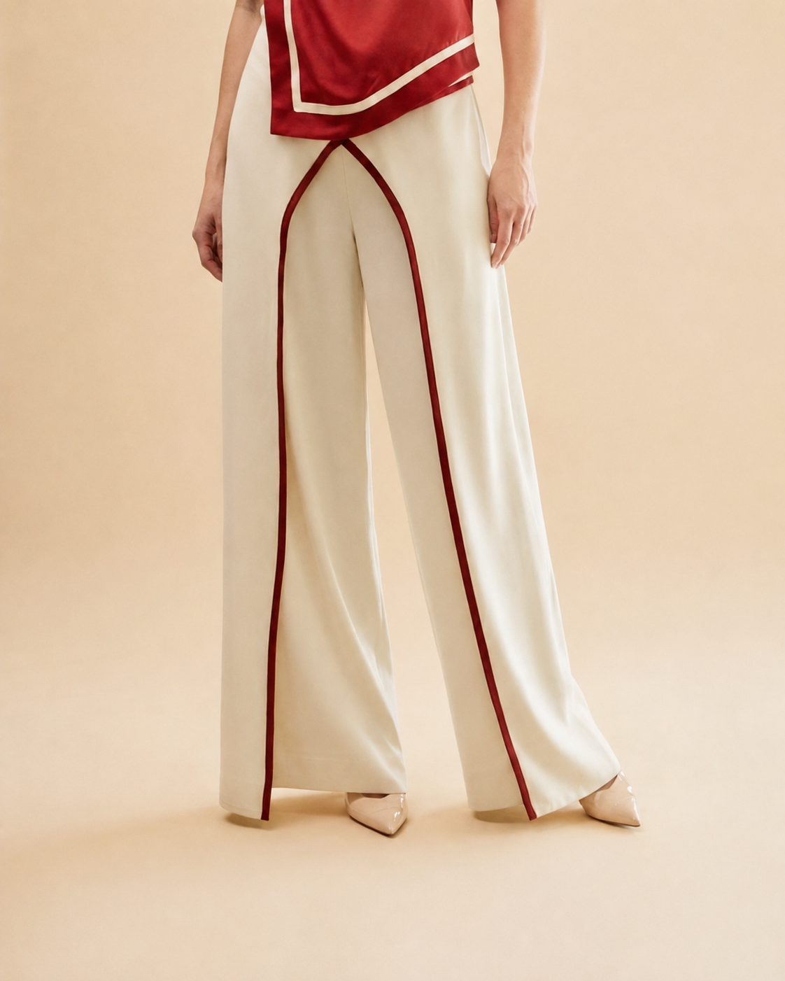 A model wearing off-white wide-leg trousers with red piping details, paired with a red top, standing against a beige stucco wall.