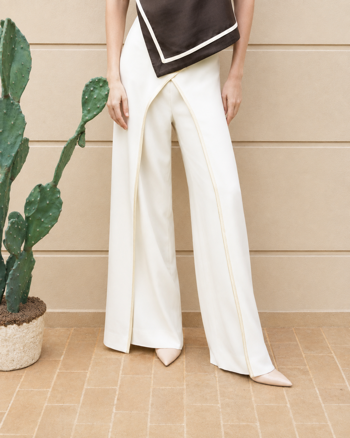 A model wearing high-waisted, wide-leg cream trousers with a draped wrap-style front and thin gold piping details, paired with a dark brown top and beige pointed-toe heels, standing in front of a beige paneled wall next to a potted cactus.