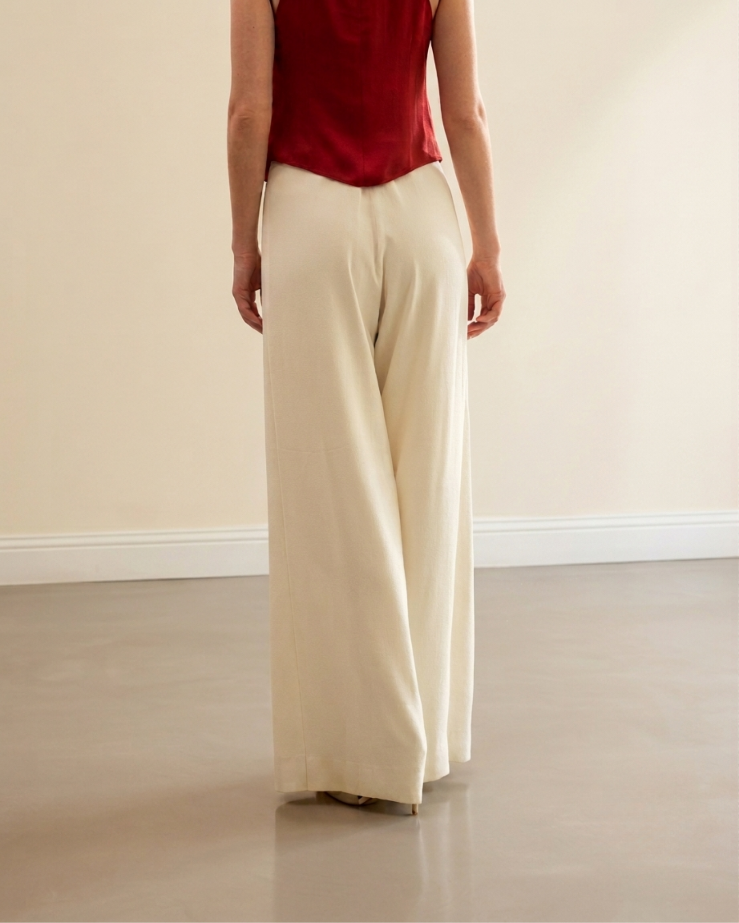 A person shown from the back wearing cream wide-leg trousers and a dark red sleeveless top, standing in a minimalist room with a beige wall and gray floor.